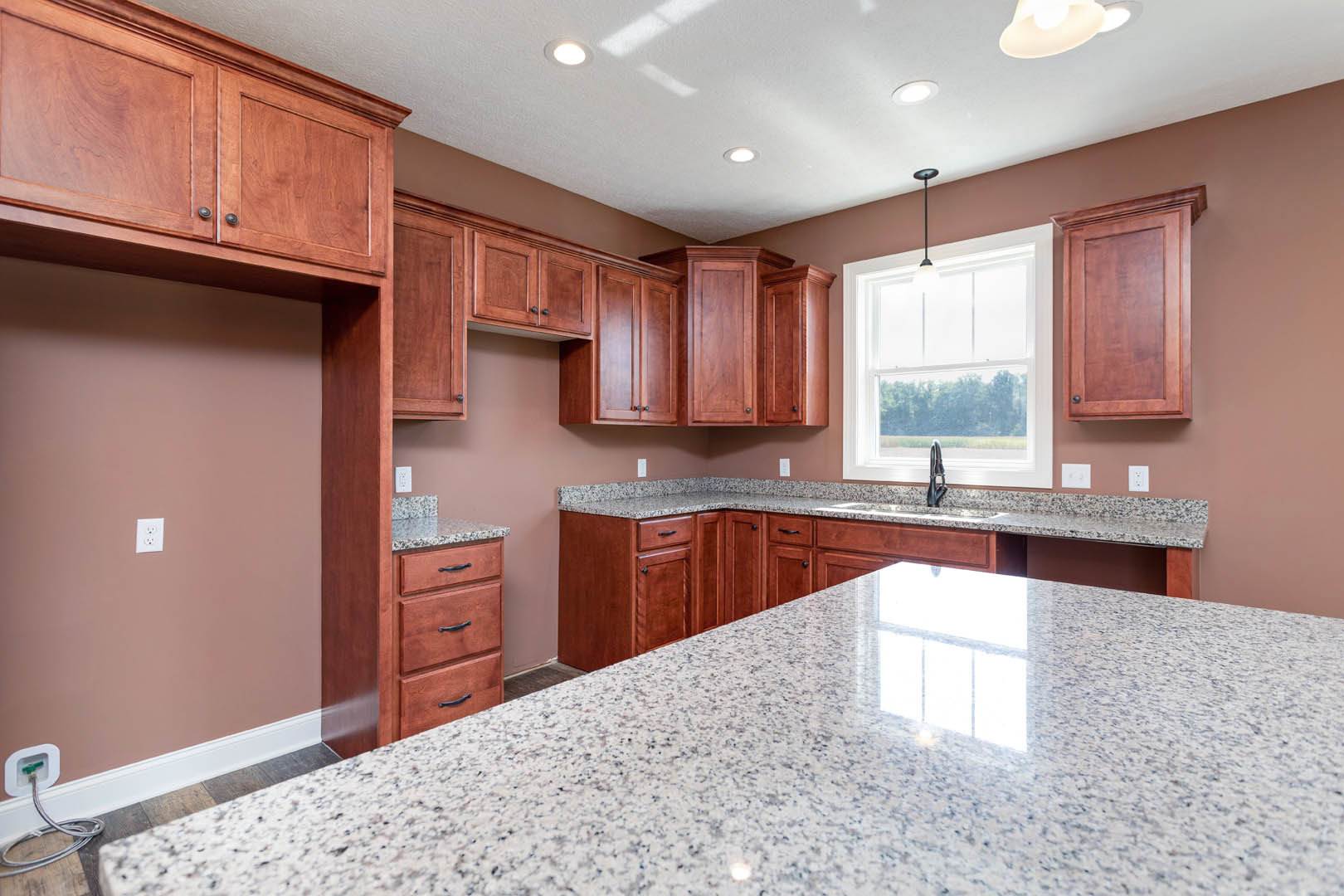 Granite countertop with marbled surface, stainless steel sink beneath window, pendant light fixture, wooden cabinetry and drawers, white backsplash