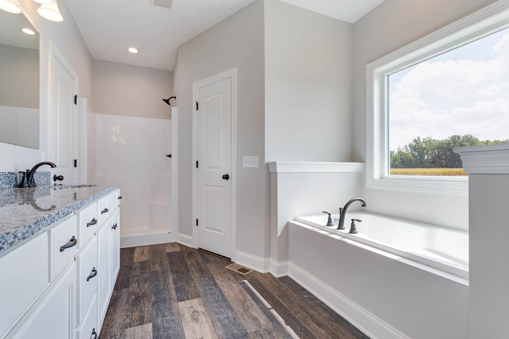 Bathroom with freestanding bathtub beneath a large window, white door with black knob, close-up of white sink and faucet, kitchen counter with white cabinetry visible in