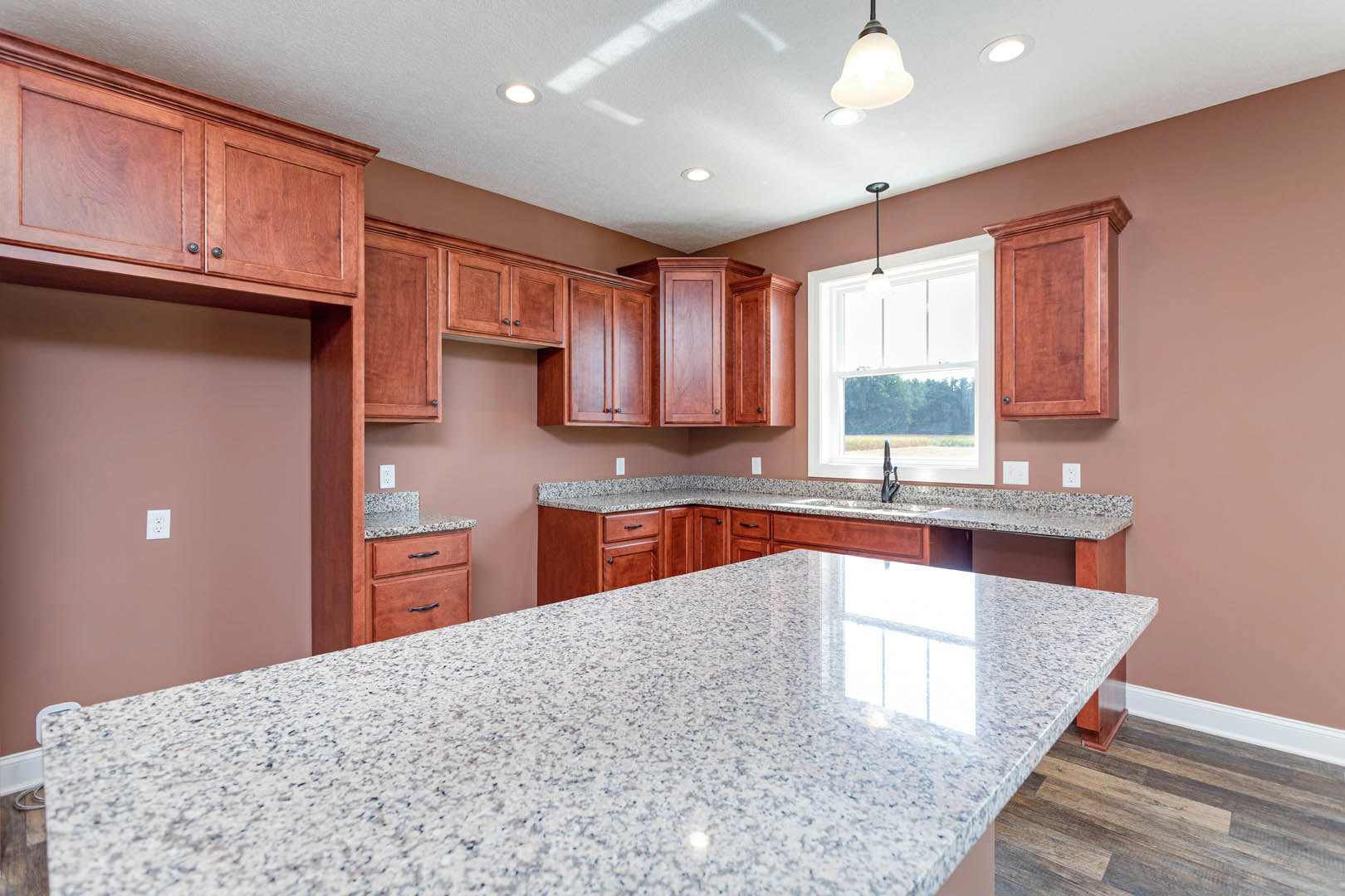 Granite countertops, wooden cabinetry, stainless steel sink, pendant light fixture above, window providing natural light in modern kitchen