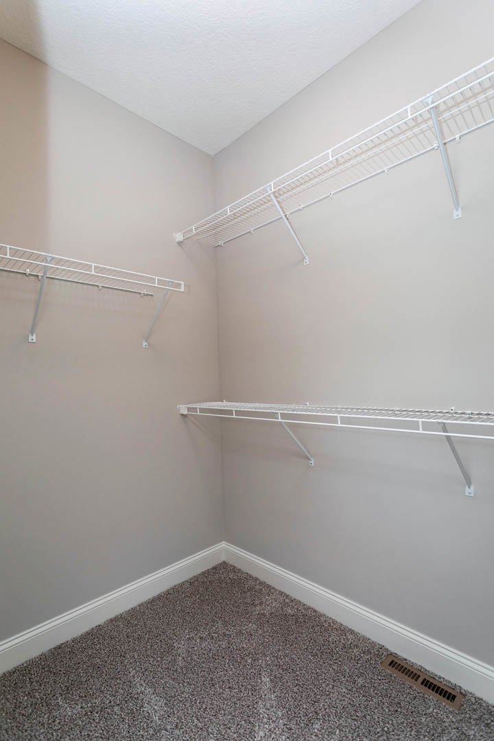 White built-in shelves on a plaster wall, carpeted corner flooring, wall vent, white metal shelf mounted above, tile flooring, and white ceiling with visible corner.