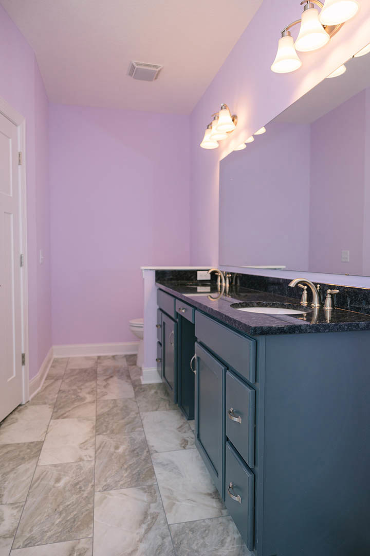 Bathroom with purple painted walls, rectangular mirror above a white sink and cabinetry, tile flooring, row of wall-mounted lights, and white door with metal handle.