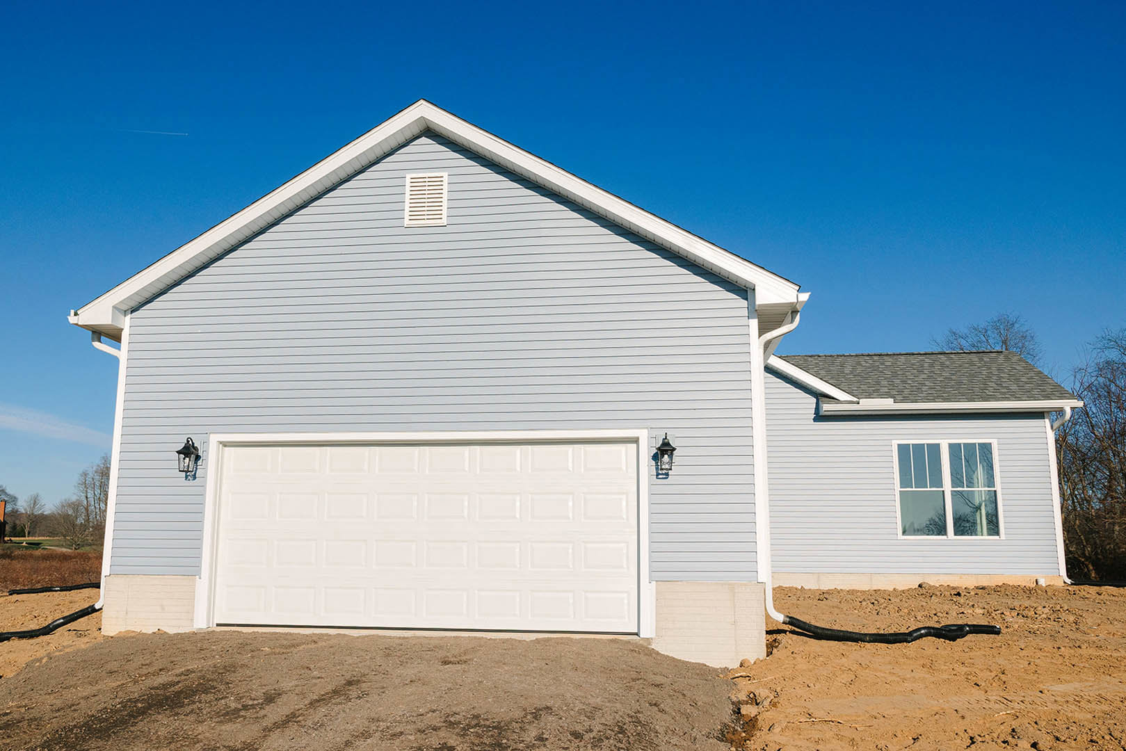 White garage door with matching trim on a custom home, gray siding exterior, single window above garage, roof vent, dirt yard with person standing near the building.