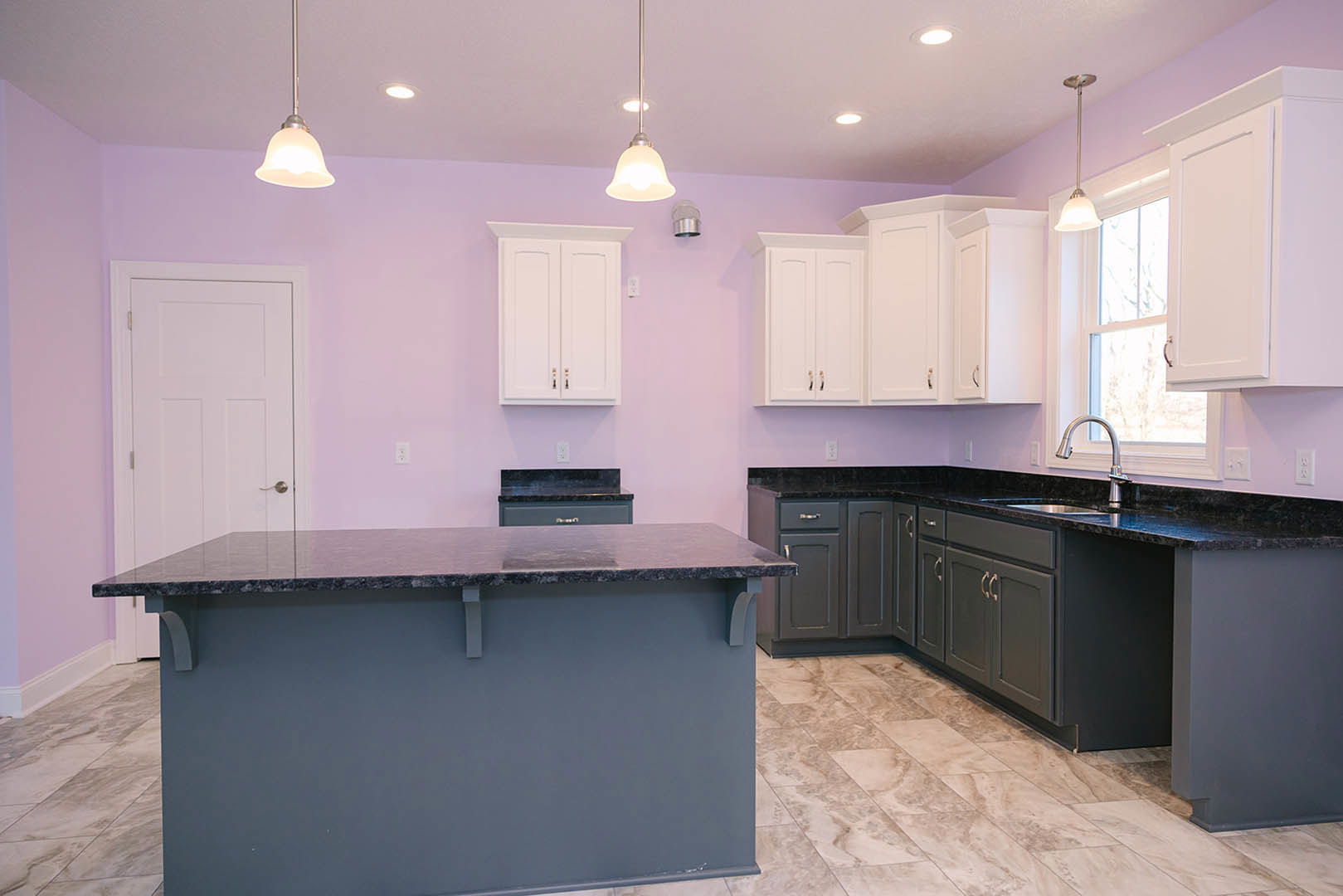 Purple kitchen walls, white cabinets and drawers with silver handles, black countertop, white door, ceiling light fixture, light-colored flooring