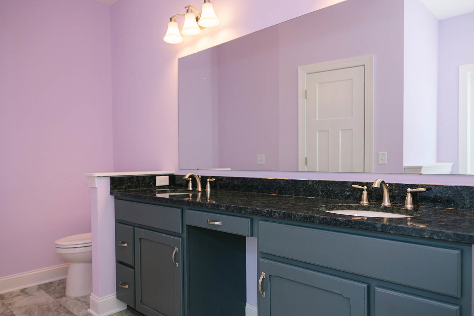 Bathroom with purple painted walls, rectangular mirror above white sink and cabinet, white door with matching frame, chrome faucet, close-up of toilet and modern light fixture.