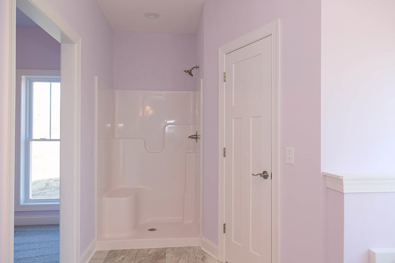 Bathroom with white tile shower featuring puzzle pattern, white door with silver handle, window, and light carpet flooring.