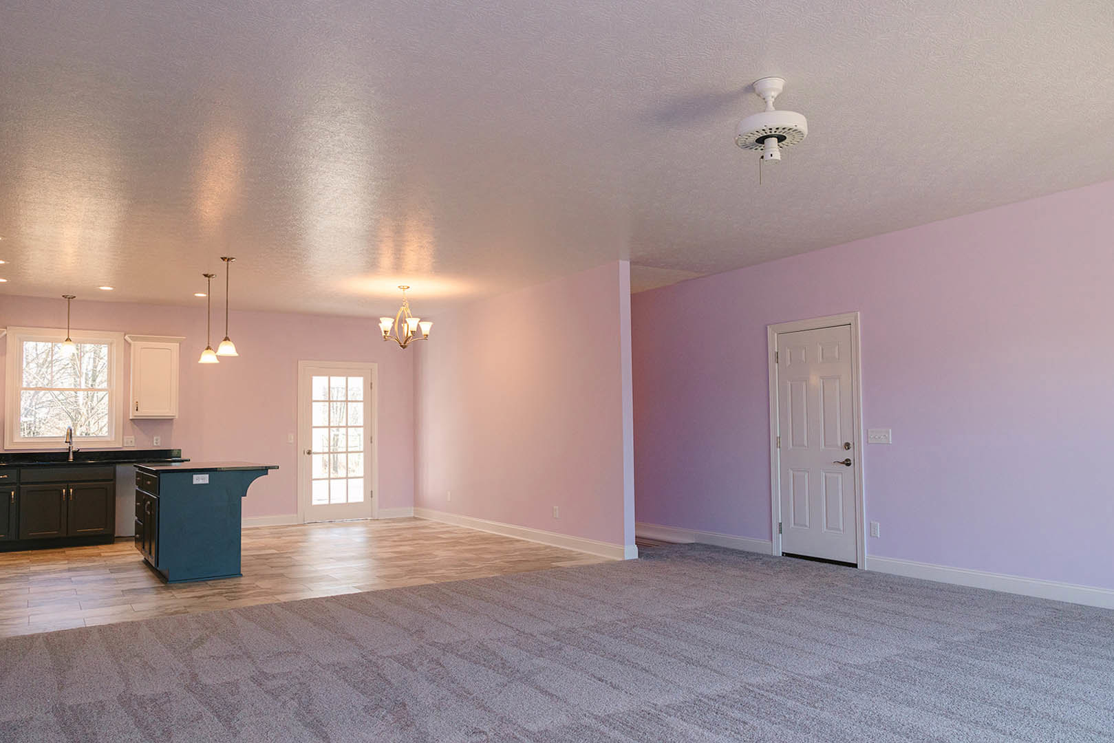 Carpeted room with a wooden table, blue desk with drawers, white door with glass panes, white ceiling fan, and adjacent kitchen sink beneath a light fixture