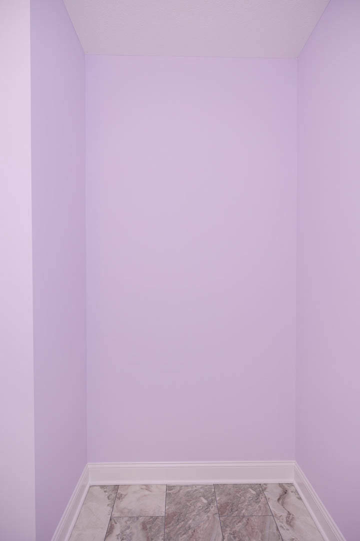 Purple accent wall adjacent to white plaster wall, tile flooring in foreground, white shelf mounted on purple wall, stone detail visible, bathroom interior.