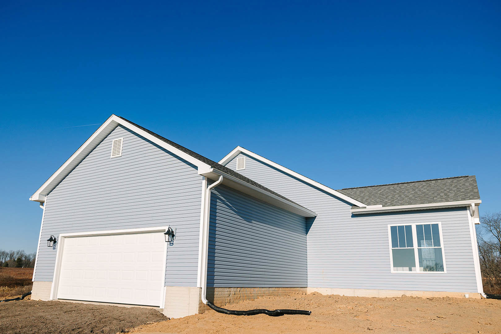 White siding house with attached garage, hose coiled near side wall, rectangular window above, vent visible, concrete driveway, blue sky overhead