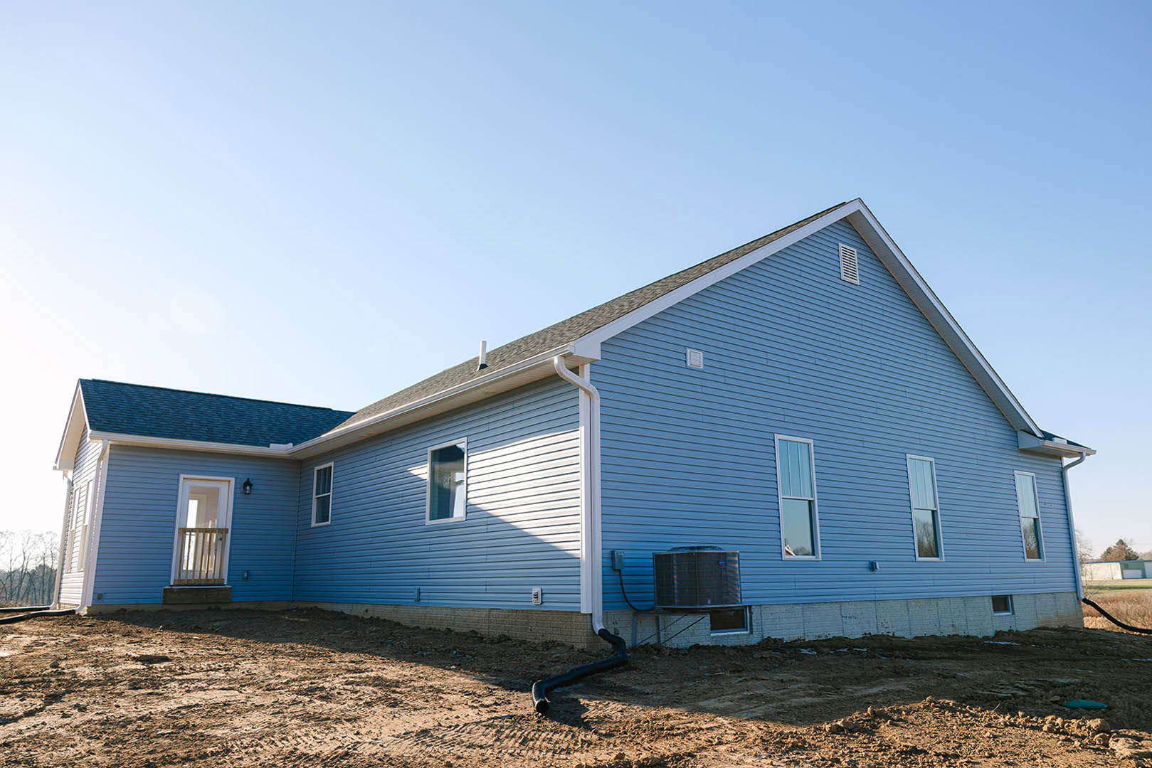 Partially built house with blue siding, exposed wooden framing, black pipe protruding near window, under clear blue sky