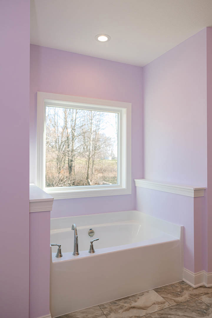 Freestanding white bathtub with chrome faucets on marble tile floor, large window overlooking green trees, smooth plaster walls