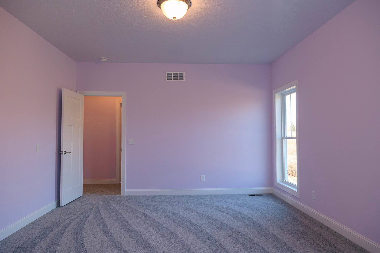 Neutral-toned carpeted room with white paneled door, painted walls, ceiling vent, recessed light, and window with trim