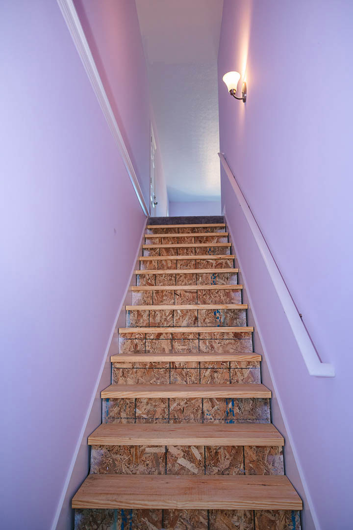 Wooden staircase with matching handrail, illuminated by a modern ceiling light fixture, purple accent wall in background, and visible wood plank finishes.