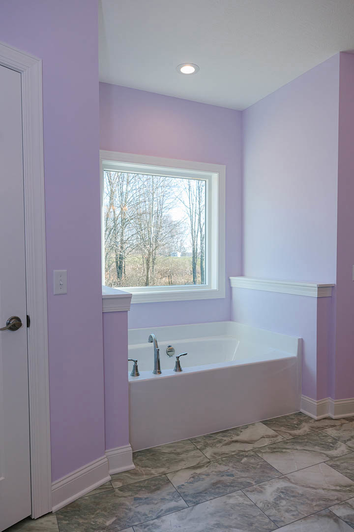 Freestanding bathtub with chrome faucets on polished marble floor, white walls with purple border, large window overlooking trees