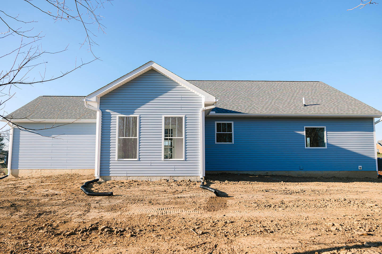 Blue siding exterior with white trim window, black pipe on dirt near foundation, clear sky in background