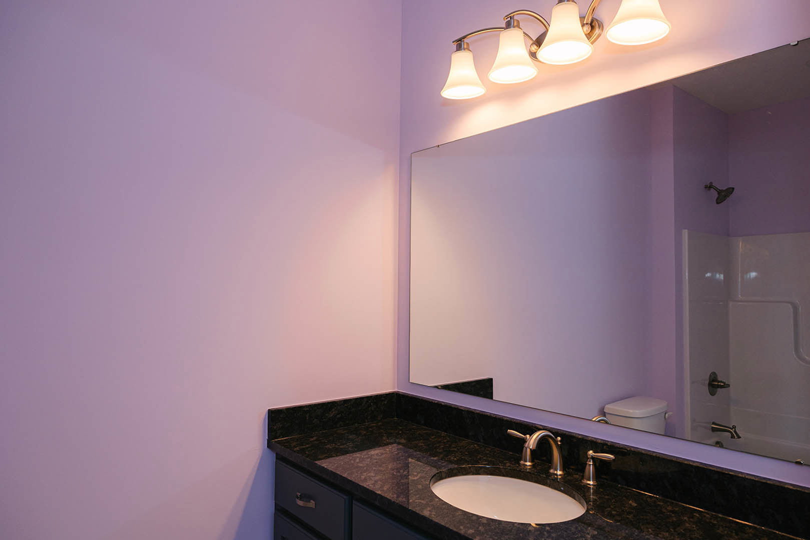 White oval sink set in a stone countertop, rectangular wall mirror above, chrome faucet, tiled backsplash, two-bulb wall sconce, partial view of glass shower enclosure, white