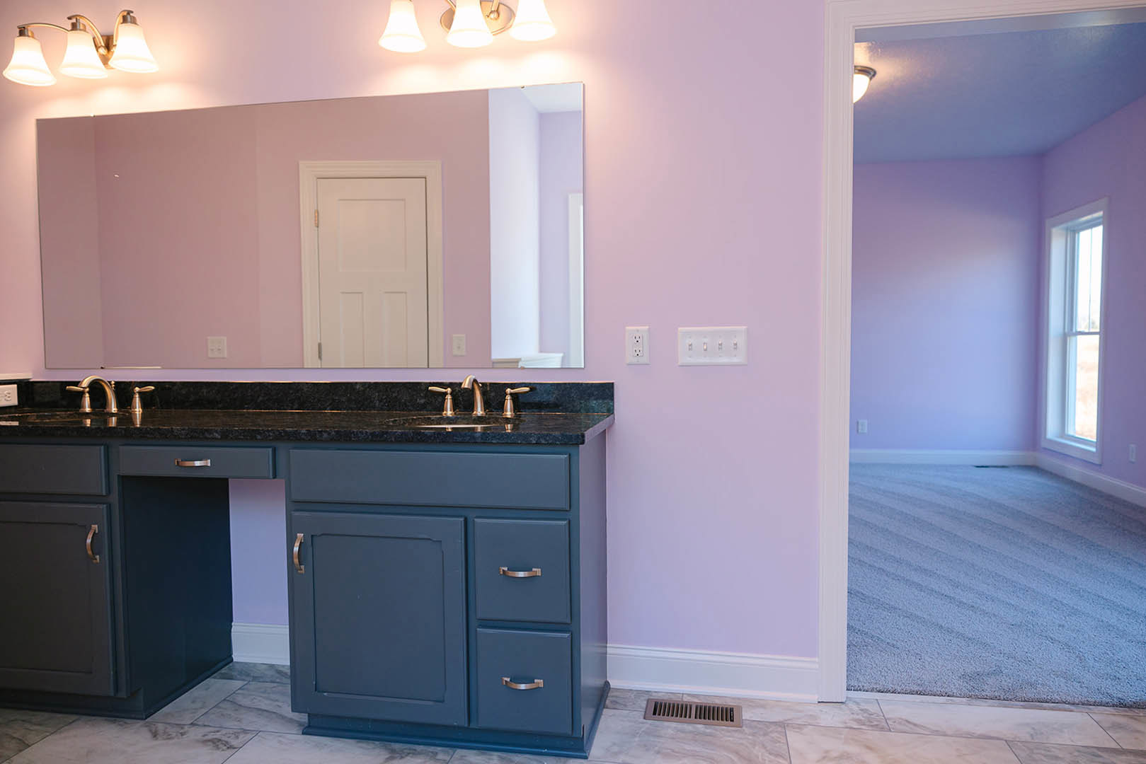 Bathroom with pink accent wall, rectangular mirror above white sink, gold hardware on white door, light-colored tile floor, white cabinetry with gold handles, and visible air vent.