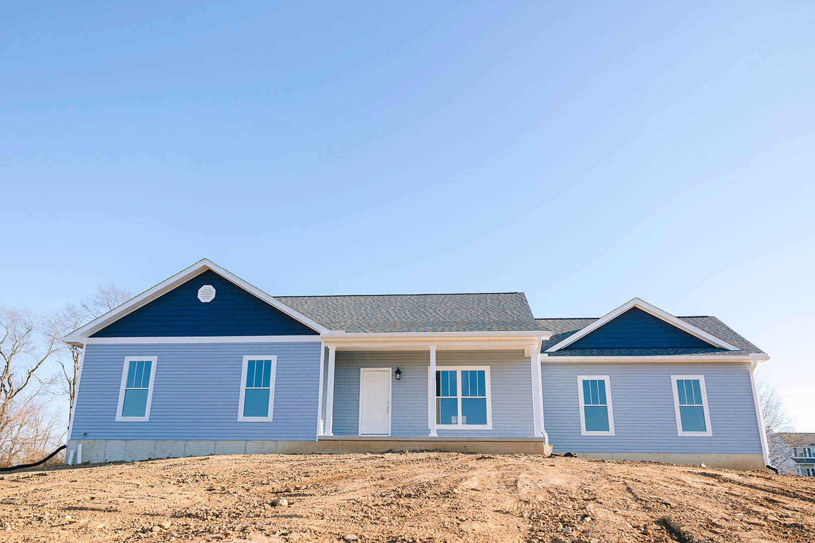 Two-story house with blue metal roof, white siding, white front door, blue shutters, and large windows; dirt yard in foreground under clear blue sky