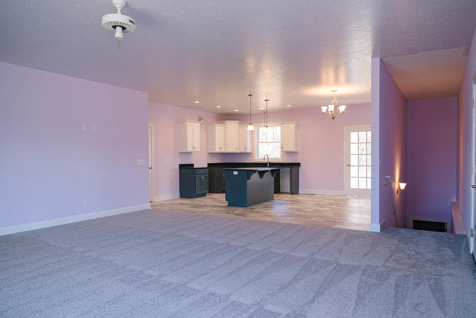 Open-concept kitchen and bar area with light wood flooring, white plaster walls, modern chandelier, and sleek black cabinetry; fireplace visible in adjoining space.