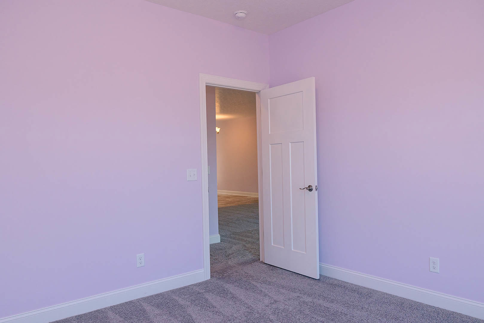 White paneled door open to a carpeted room with light purple walls, wall-mounted light fixture, and plaster finish