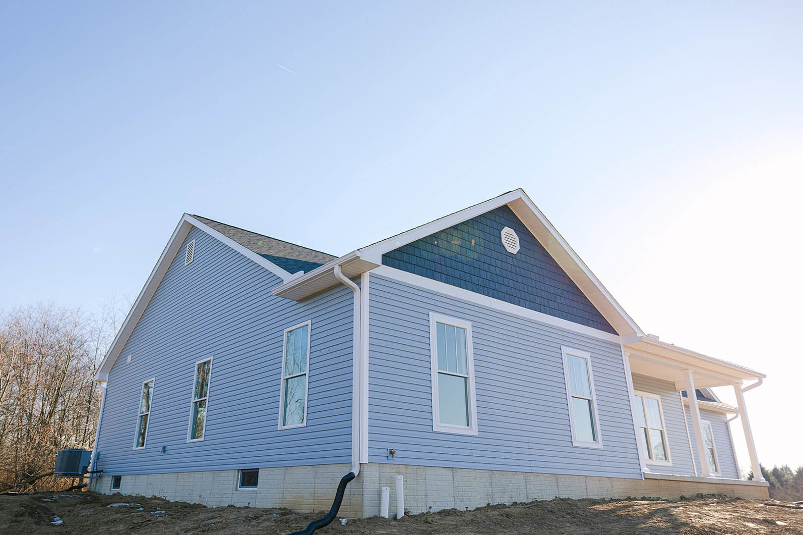 Two-story house under construction with blue siding, white-framed window, exposed roof trusses, leafless tree in foreground, clear blue sky overhead