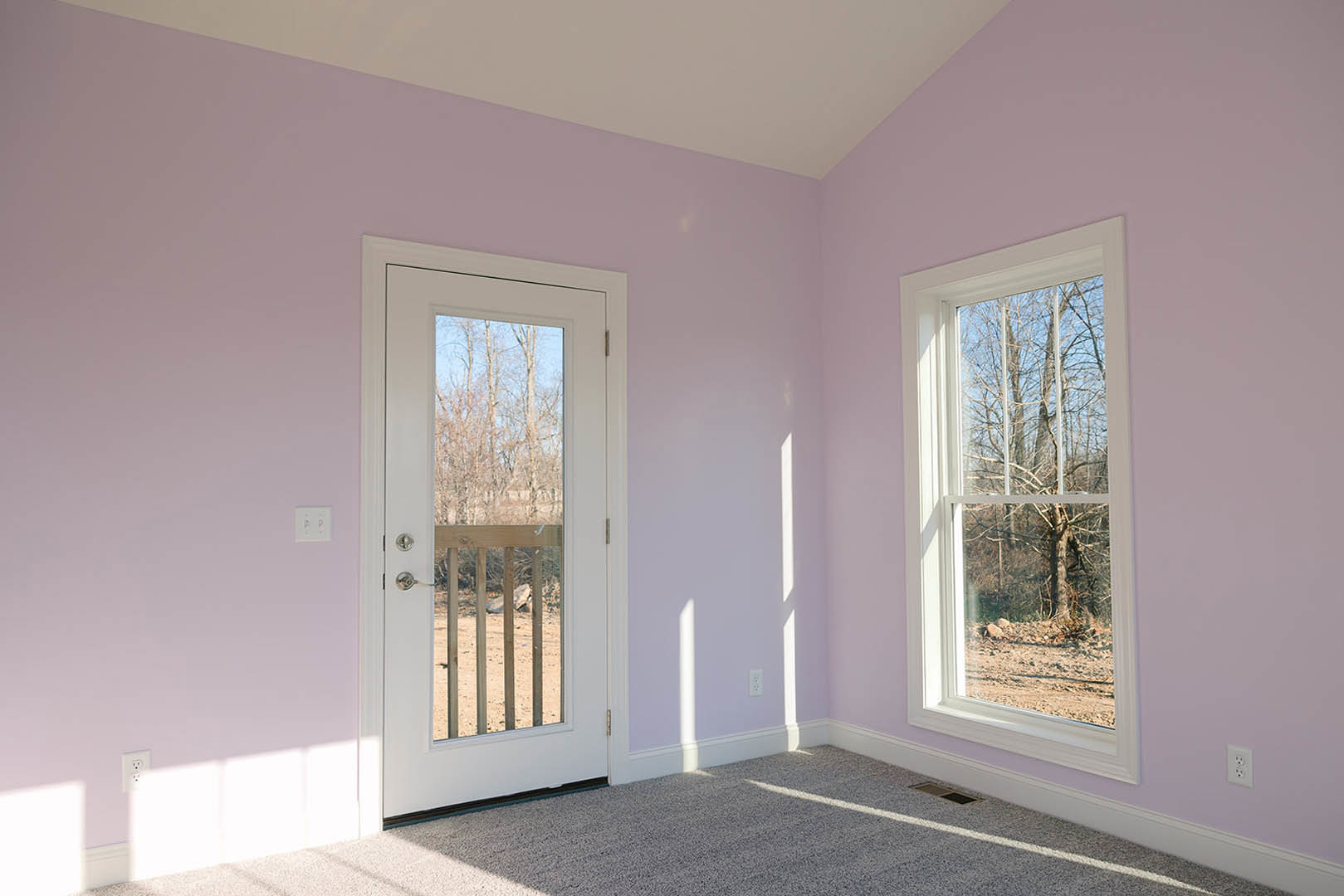 Bright room with white walls, large windows showing green trees, wood door, light carpet flooring, white wall outlet with black switches, and baseboard molding