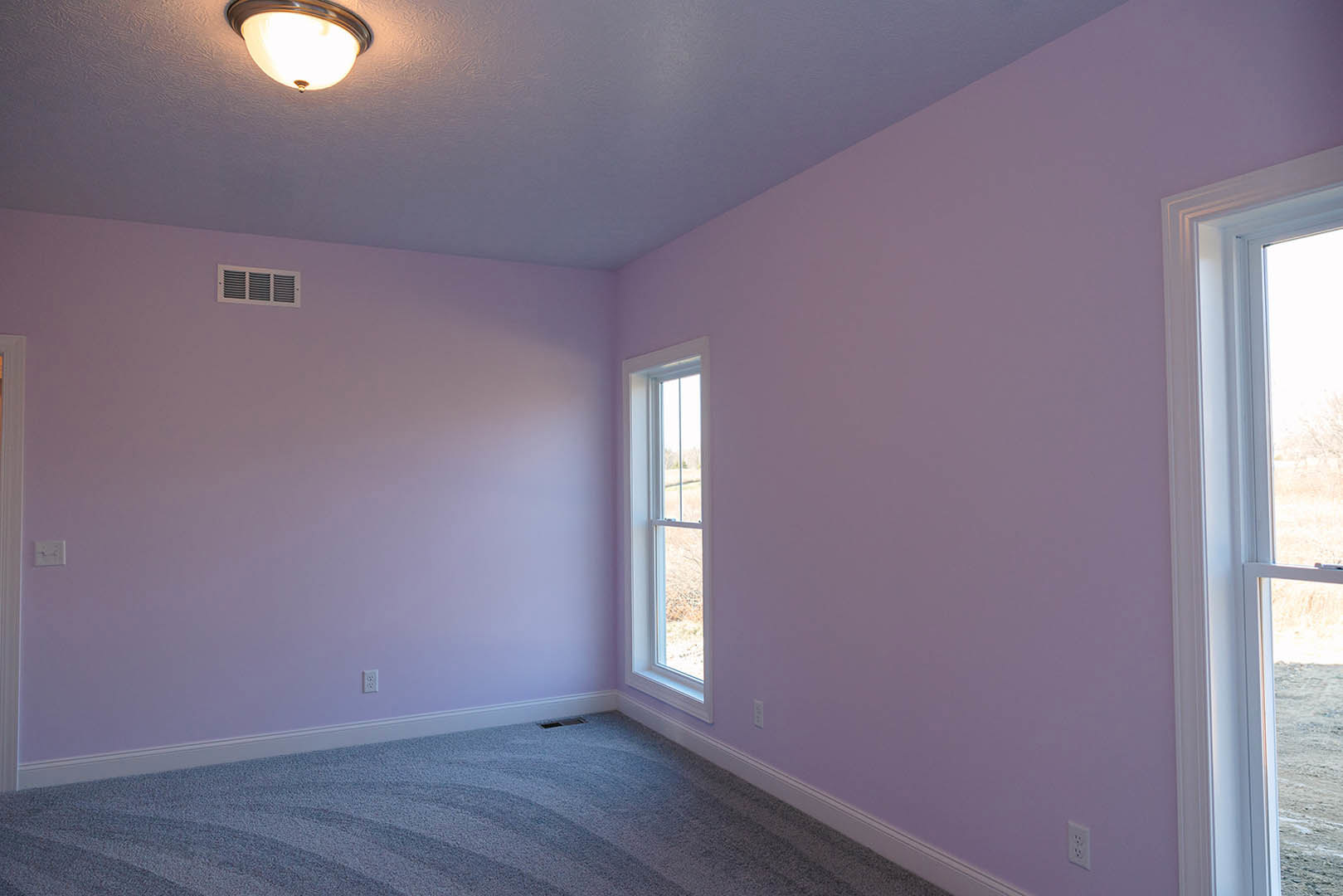 Ceiling-mounted light fixture above hardwood flooring, white plaster walls, large window with view of grassy field, rectangular wall vent