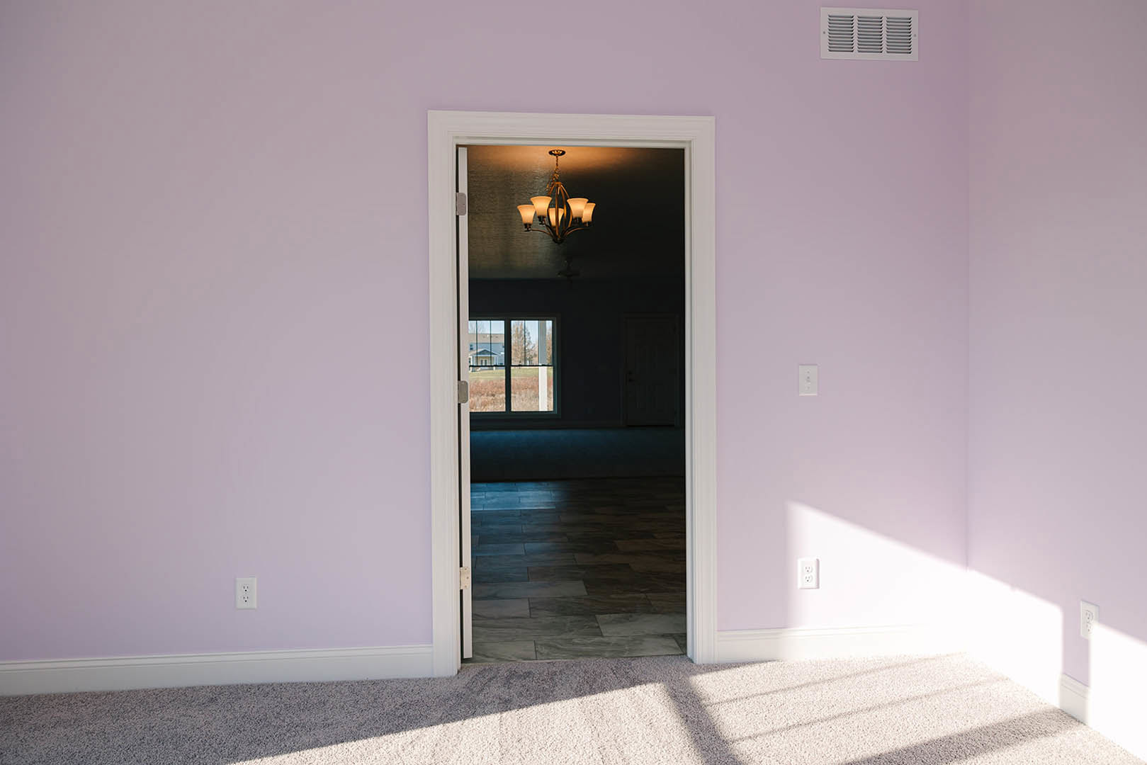 Open white door in a room with dark tiled floor, four-light ceiling fixture, window overlooking a field, visible carpet and vent details