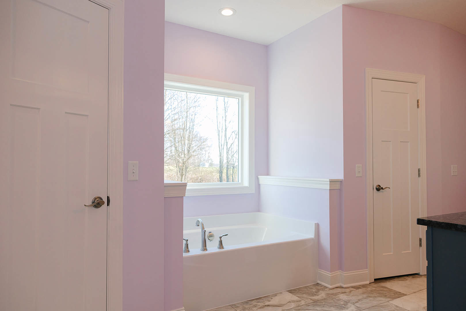 White bathtub with chrome faucets against a white wall, large window overlooking trees, blue accent wall, and partial view of a door