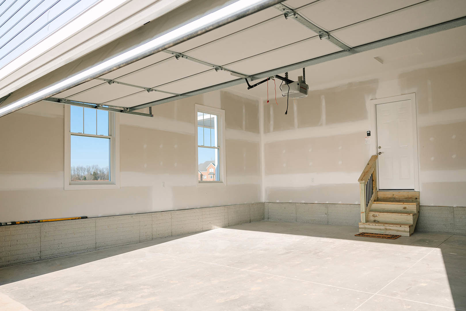 Open room with polished concrete floor, wooden staircase with black metal railing, white garage door, plaster walls, and large window showing clear blue sky.