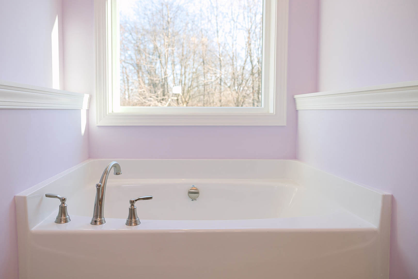 Freestanding white bathtub with chrome faucet beneath a large window overlooking leafy trees, set against light-colored bathroom walls