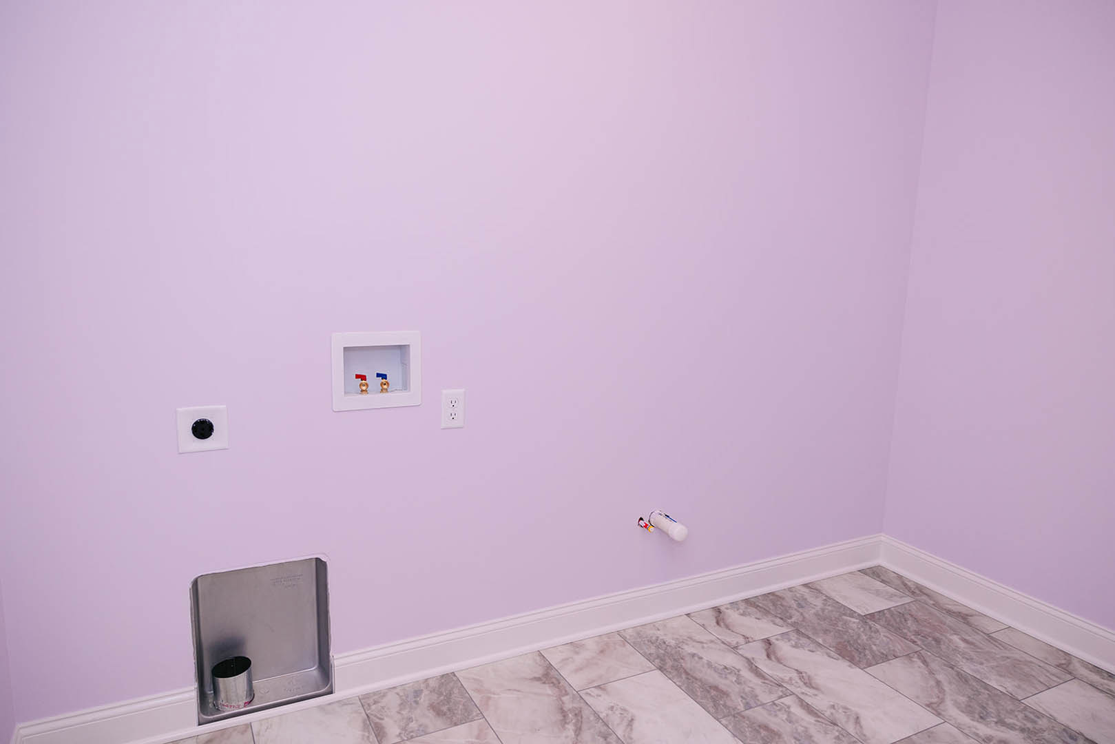 Purple plaster wall and tile floor in a bathroom, featuring a white square box with a white frame and red and blue valve, metal can in a recessed area, white bottle with blue and