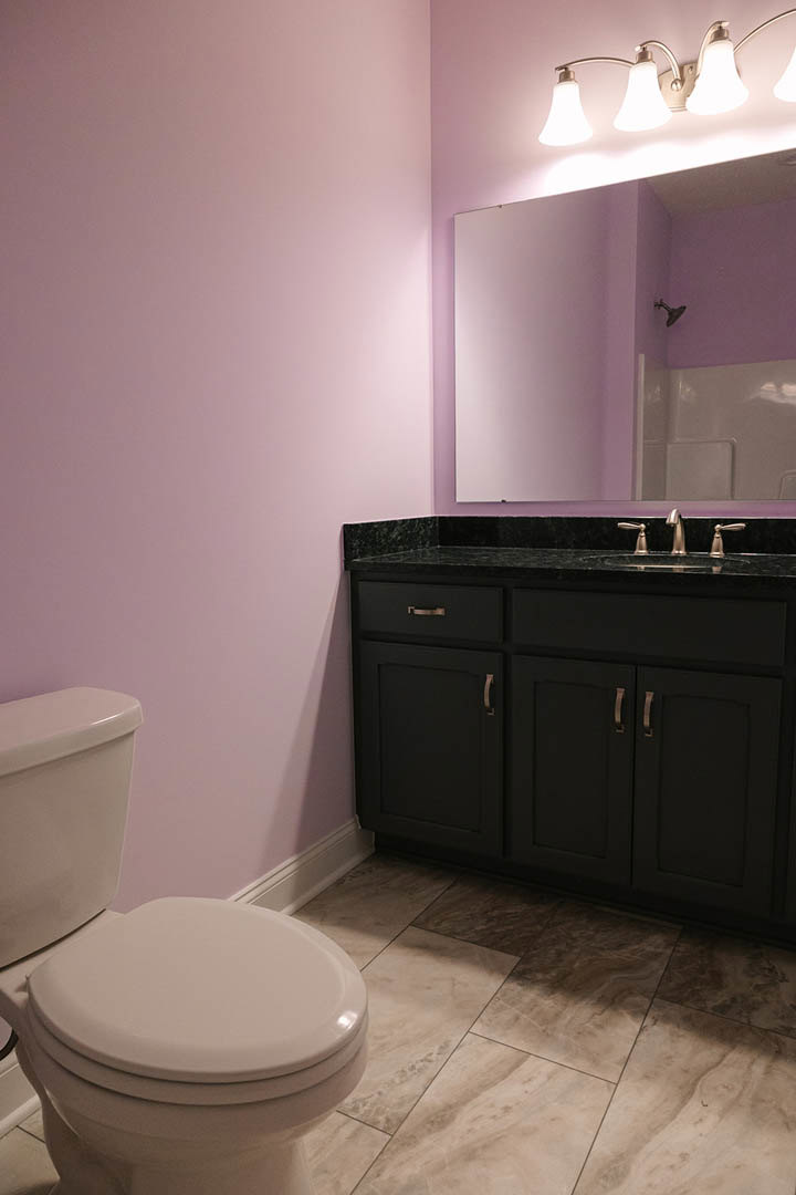 Bathroom with purple painted walls, white toilet, black vanity with marble countertop, tile flooring, modern light fixture, and glass shower enclosure