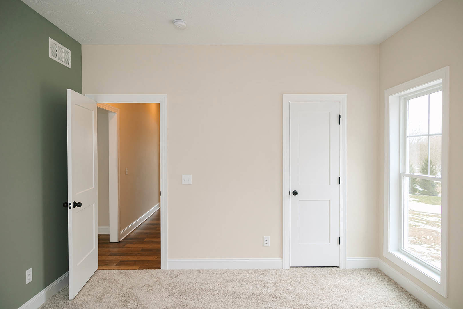 Bedroom with plush beige carpet, white paneled doors featuring black hardware, wall vent, and window overlooking snowy landscape