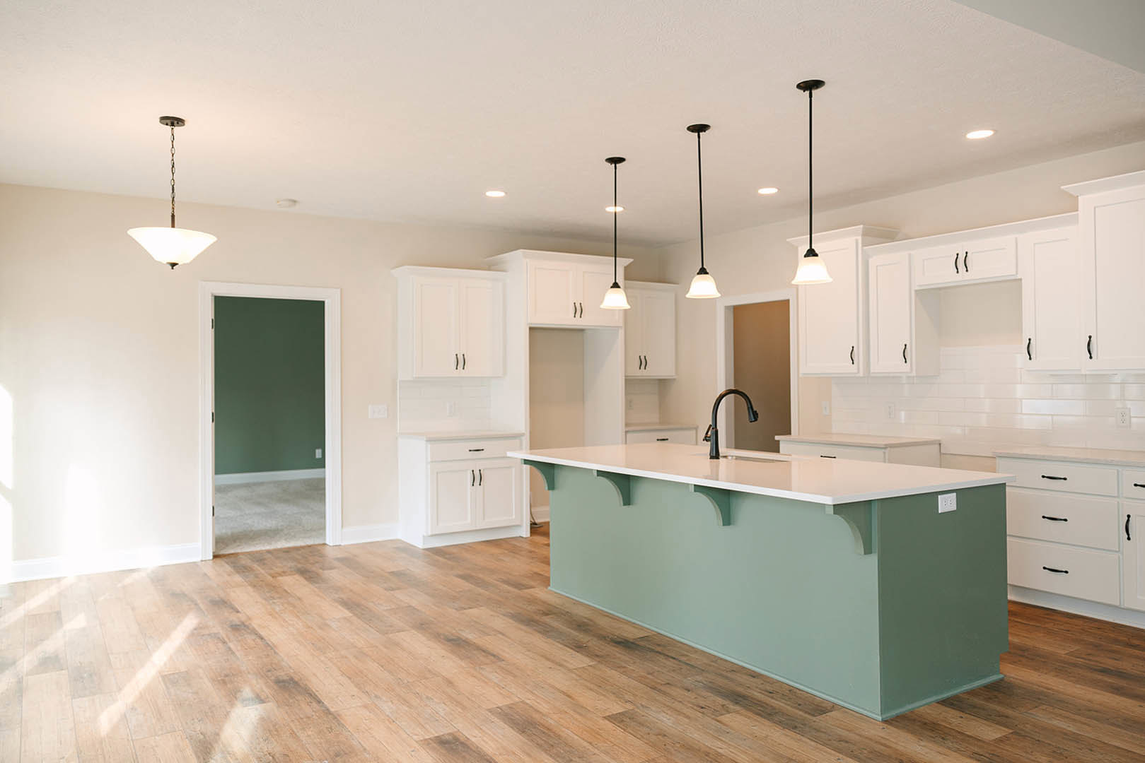 Kitchen with white shaker cabinets, central island featuring white countertop, black pendant lamp overhead, green accent wall with white trim, wood flooring, stainless steel sink