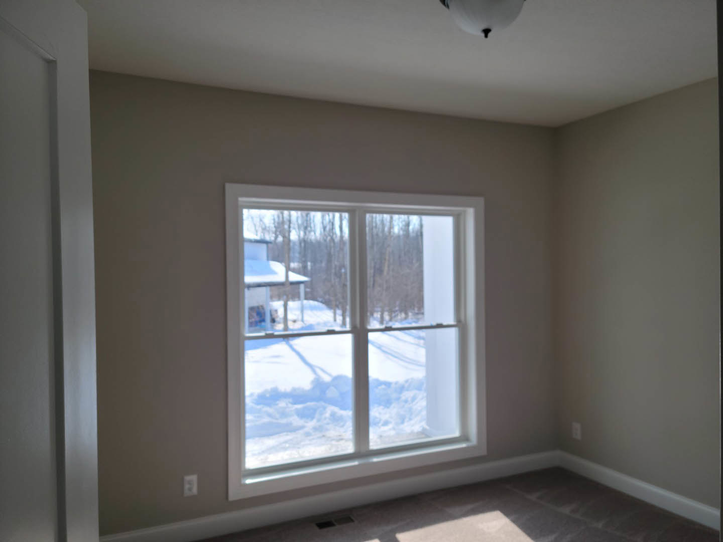 Large window with white trim overlooking snow-covered yard, soft natural light filling neutral-toned room, partially visible wall and ceiling, modern light fixture blurred in