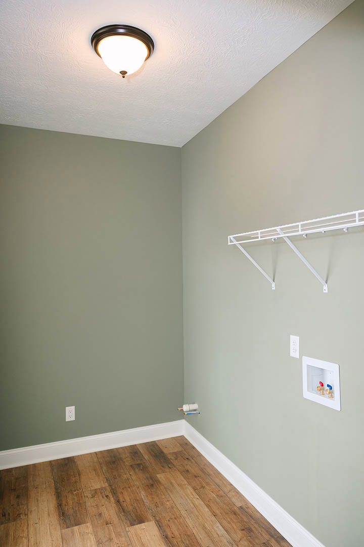 Wood flooring with white baseboards, white metal wall shelf featuring a ladder, brass faucets on a white box, ceiling light fixture, plaster walls.