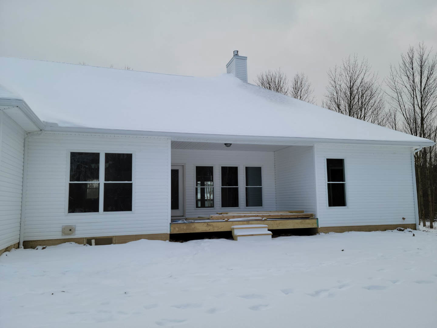 Two-story house with gray siding, white trim, and chimney, surrounded by snow-covered ground; steps lead to front entrance, large windows reflect bare tree branches