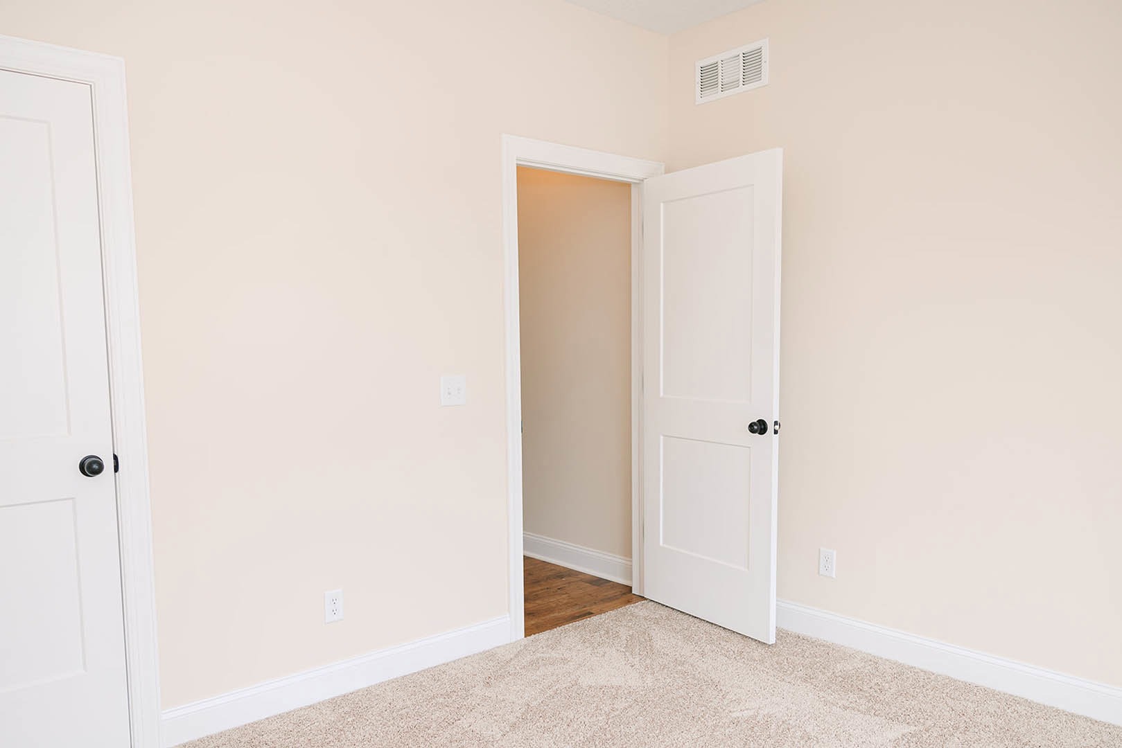 White paneled door with black knob set in a white wall, white baseboard trim, carpeted floor, and a white vent visible nearby