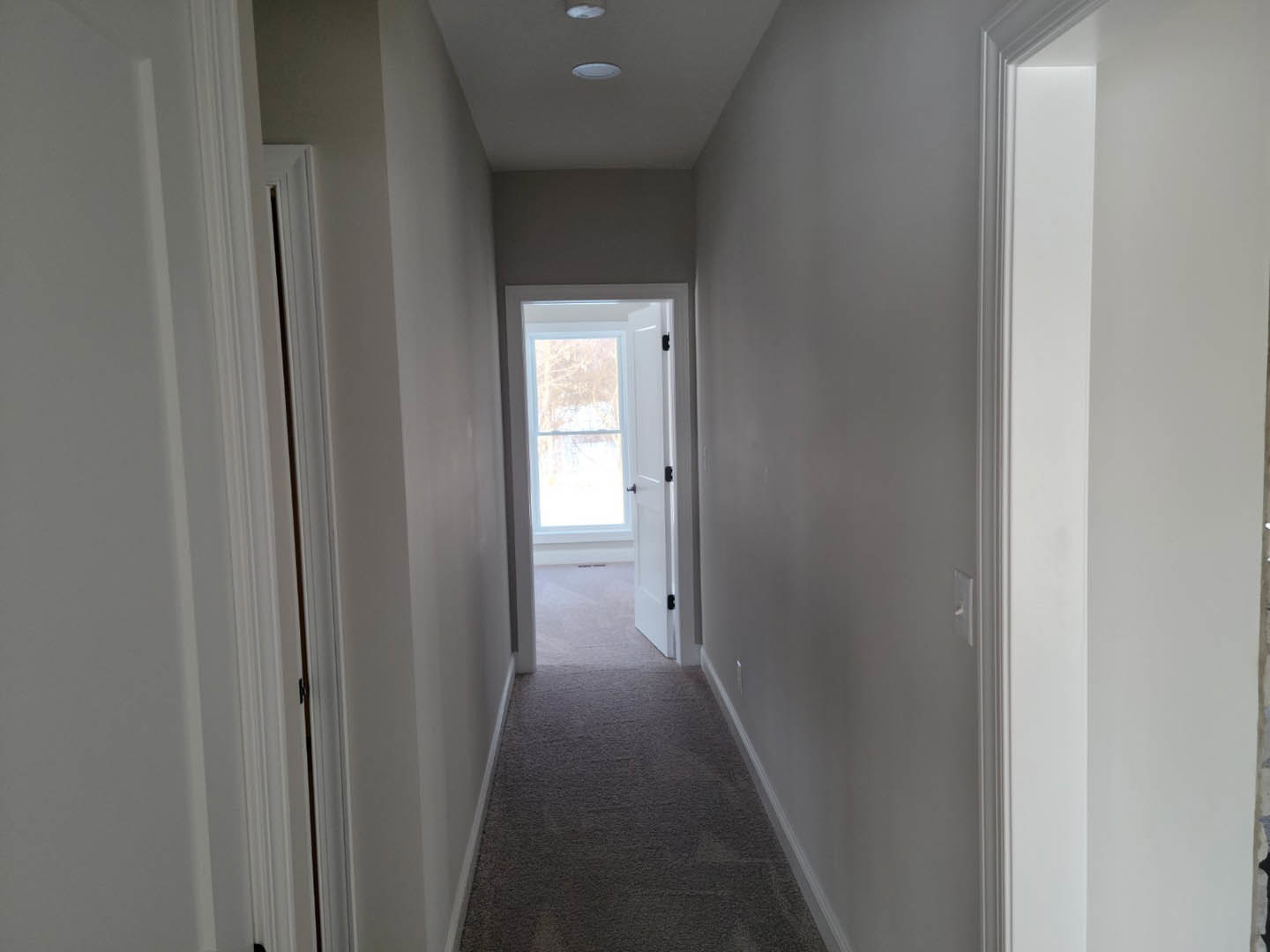 Hallway with light-colored plaster walls, white door featuring black handle, open to reveal window framing snowy landscape, wood flooring, and white ceiling