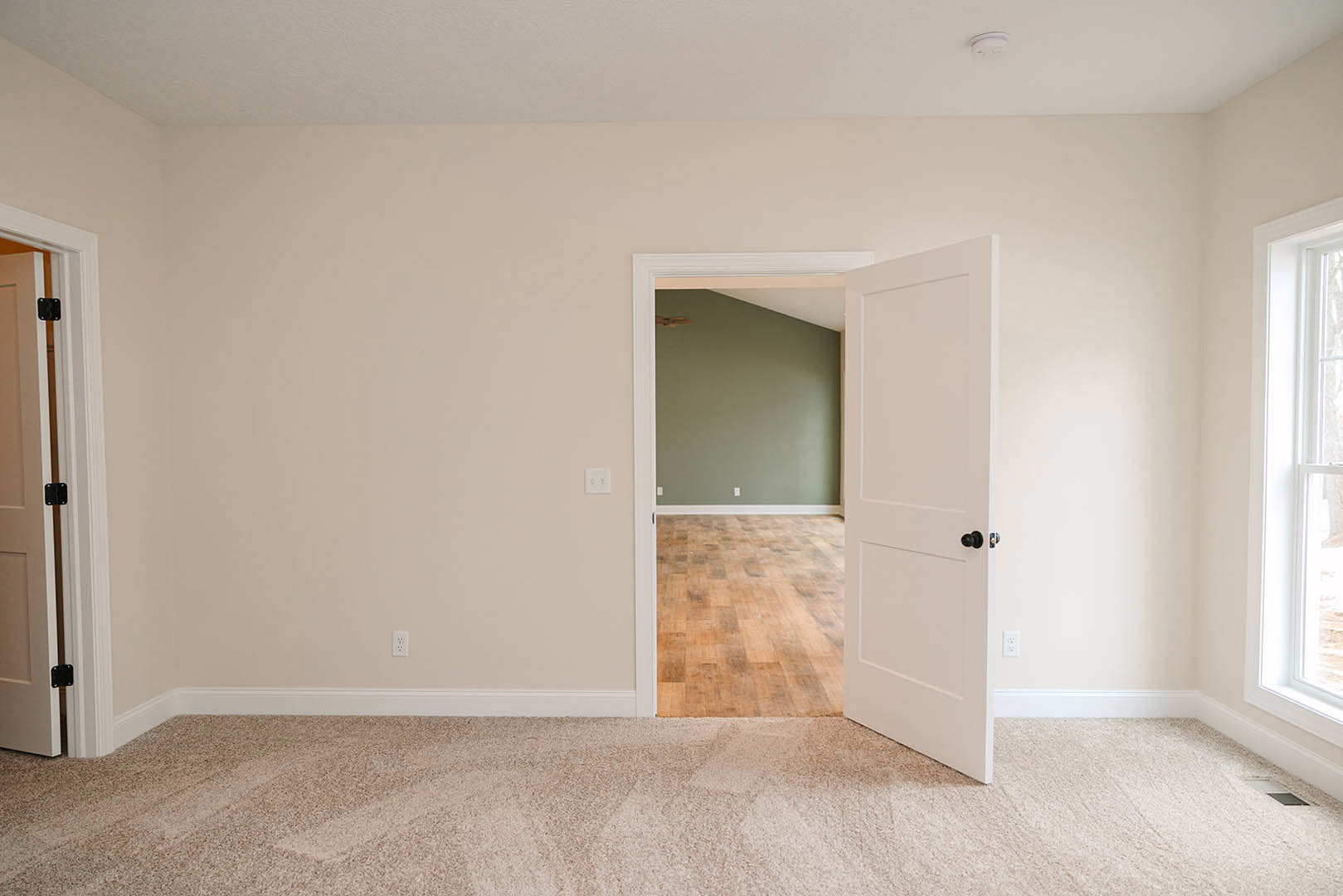Open white door with brushed metal hinge, leading from room with light wood laminate flooring and white baseboards; partial view of window and beige carpet in adjacent space.