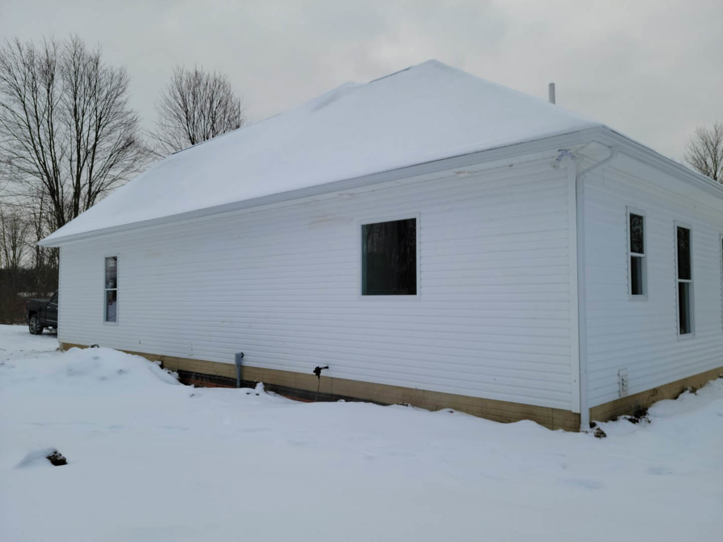 White house with snow-covered roof and ground, leafless tree in front yard, black truck parked near window, person holding hose beside building, winter sky overhead