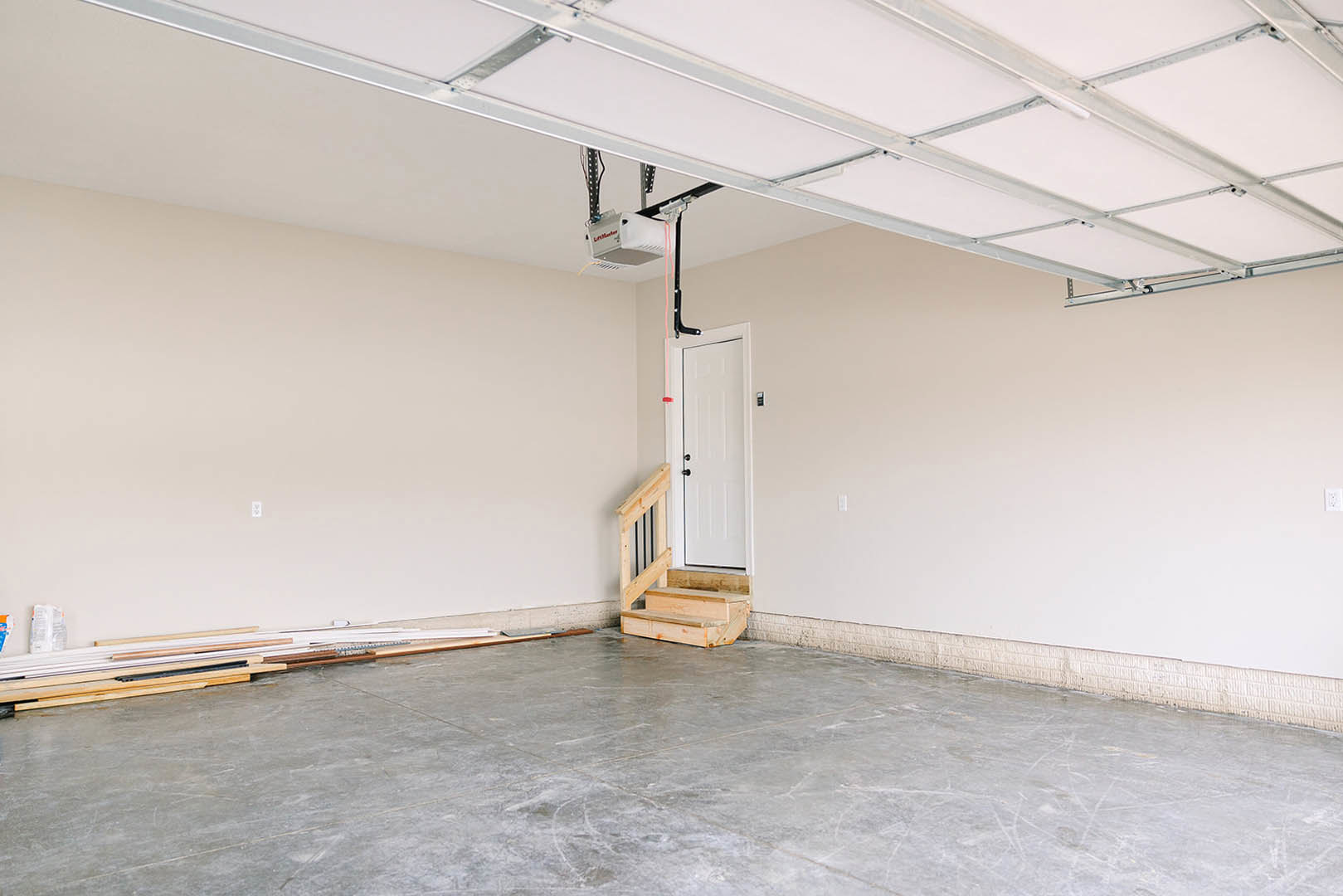 White plaster wall and door with black knobs, concrete and tile flooring, garage door opener mounted on ceiling, white food bag and white box in empty room.