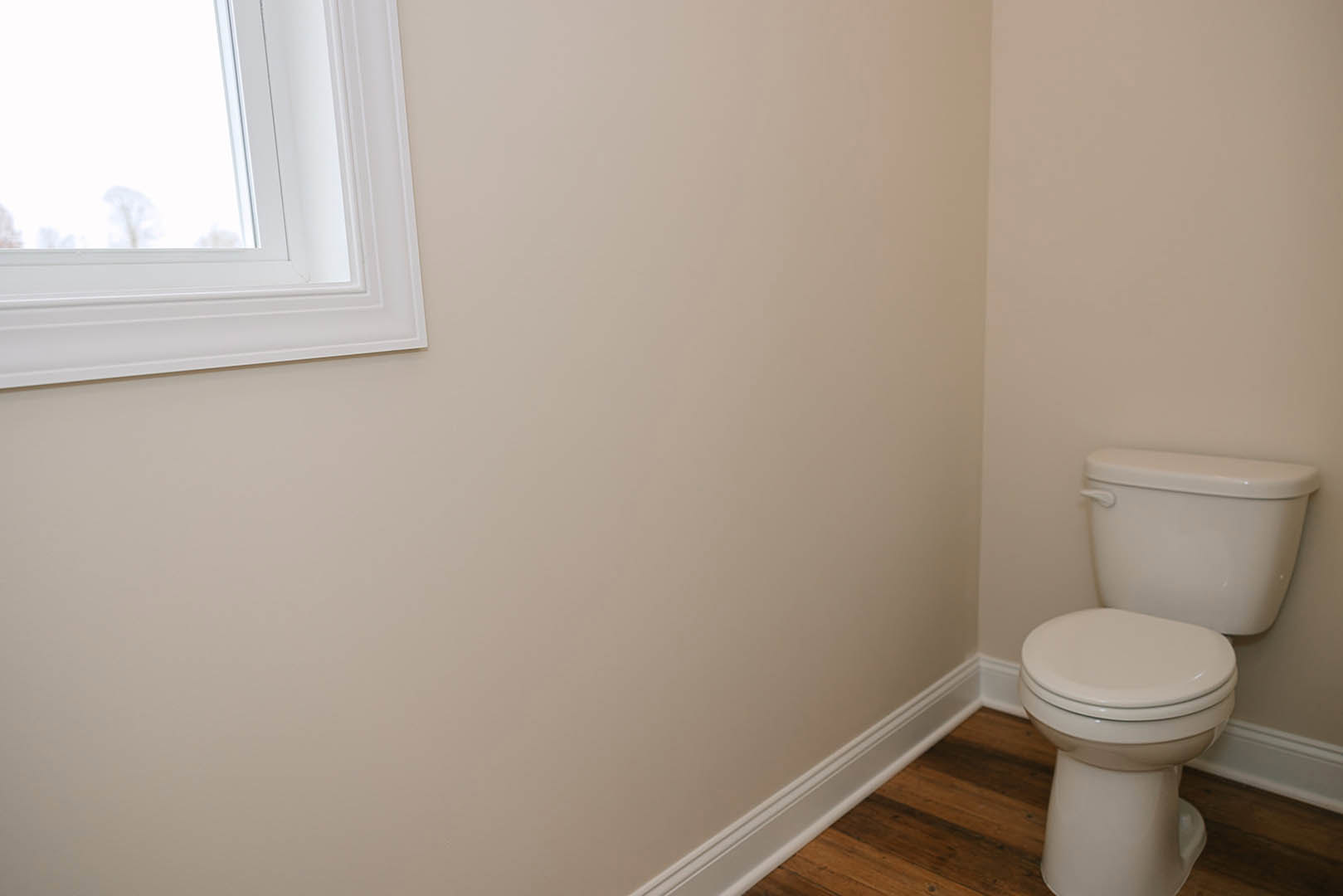 White toilet with closed seat on wood flooring, adjacent to a plaster wall and window, in a modern bathroom interior