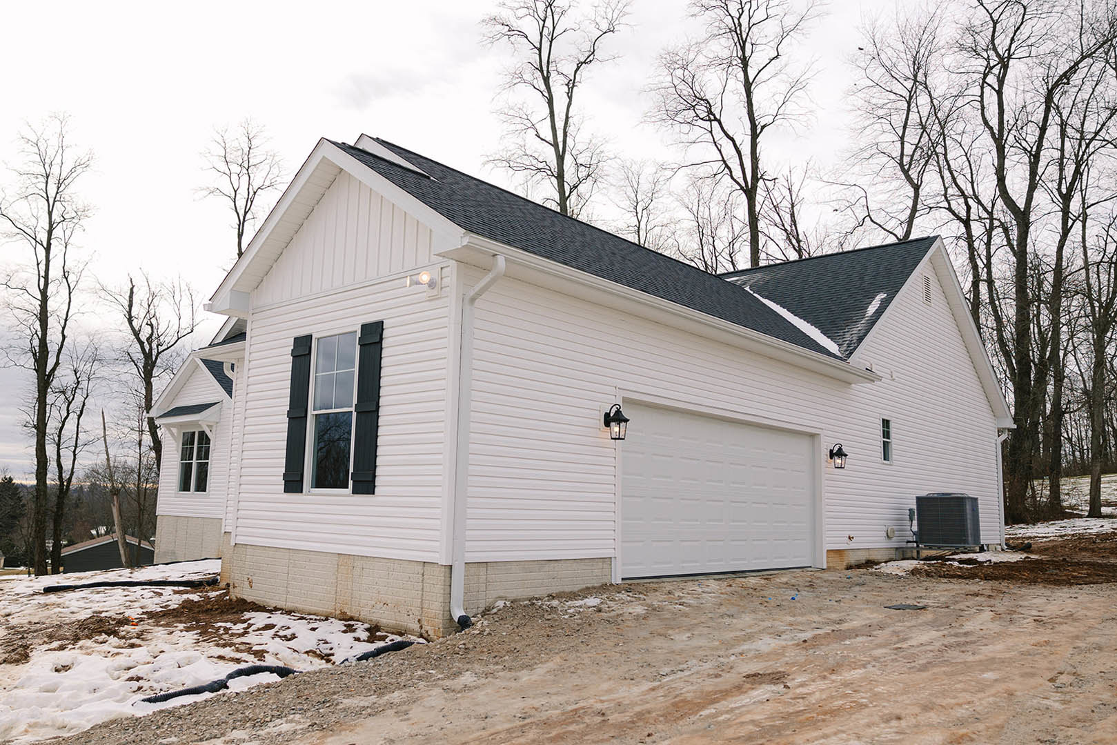White house with attached garage, driveway partially covered in snow, leafless tree in front yard, close-up window, large metal box on pallet, black pipe lying in snow.