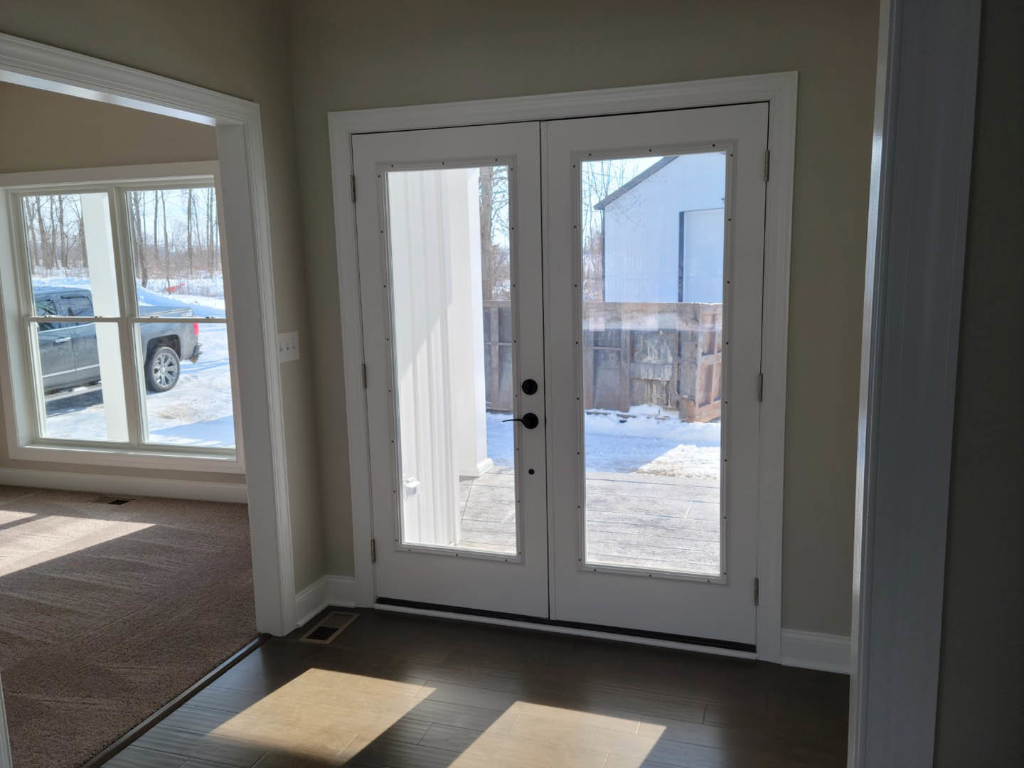 Double door with clear glass panes, white trim, and carpeted flooring inside a residential entryway, snowy driveway and parked truck visible through window.