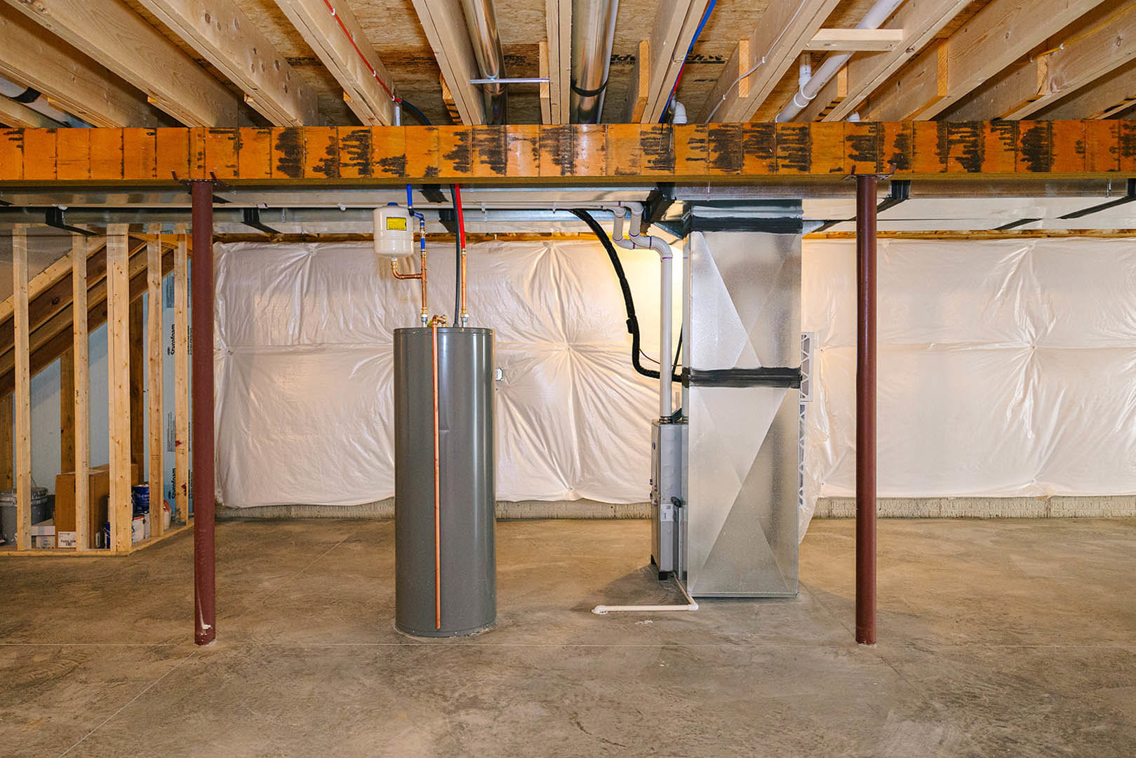 Basement utility room with exposed beams, grey metal cylinder heater, red and blue pipes, white container with blue lid, and unfinished concrete floor.
