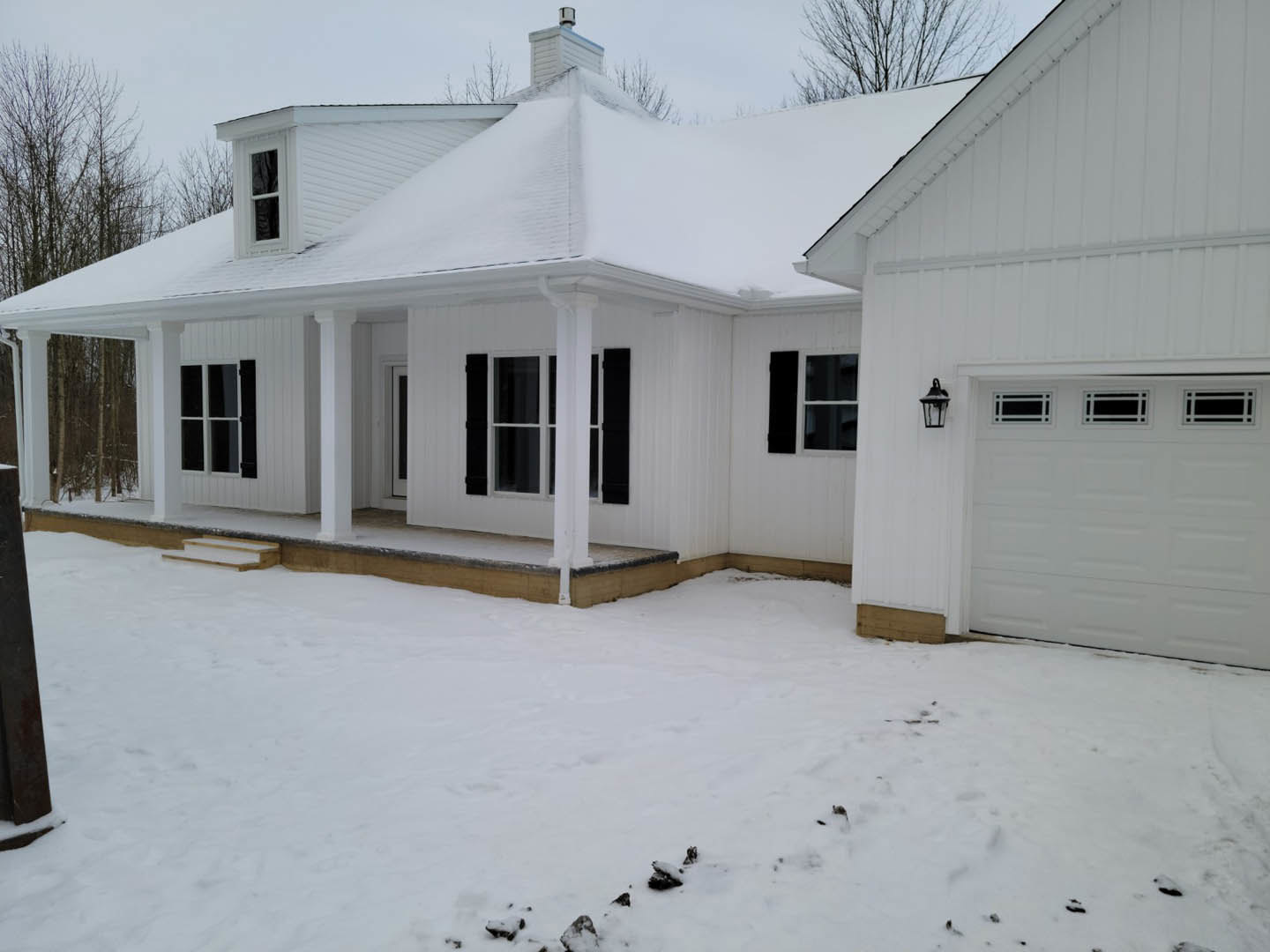 White siding house with attached garage, white-framed windows, and covered porch, surrounded by snow-covered ground under a winter sky