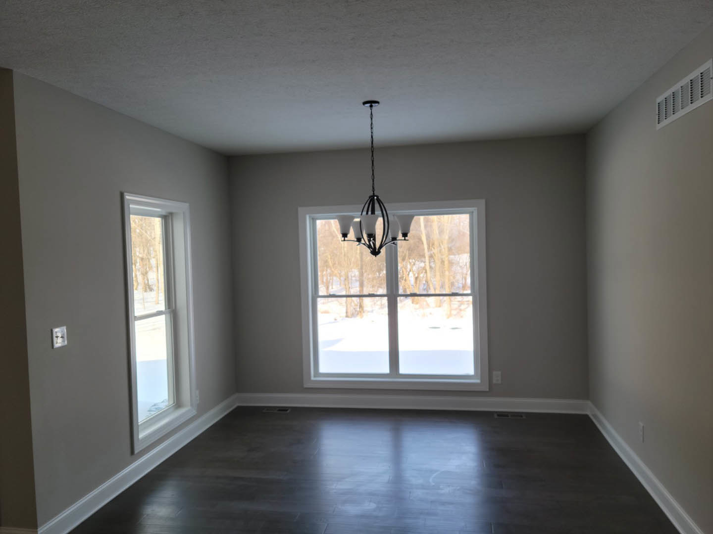 Chandelier hanging from white ceiling above dark wood laminate flooring, large windows with snowy landscape view, plaster walls, wall vent visible