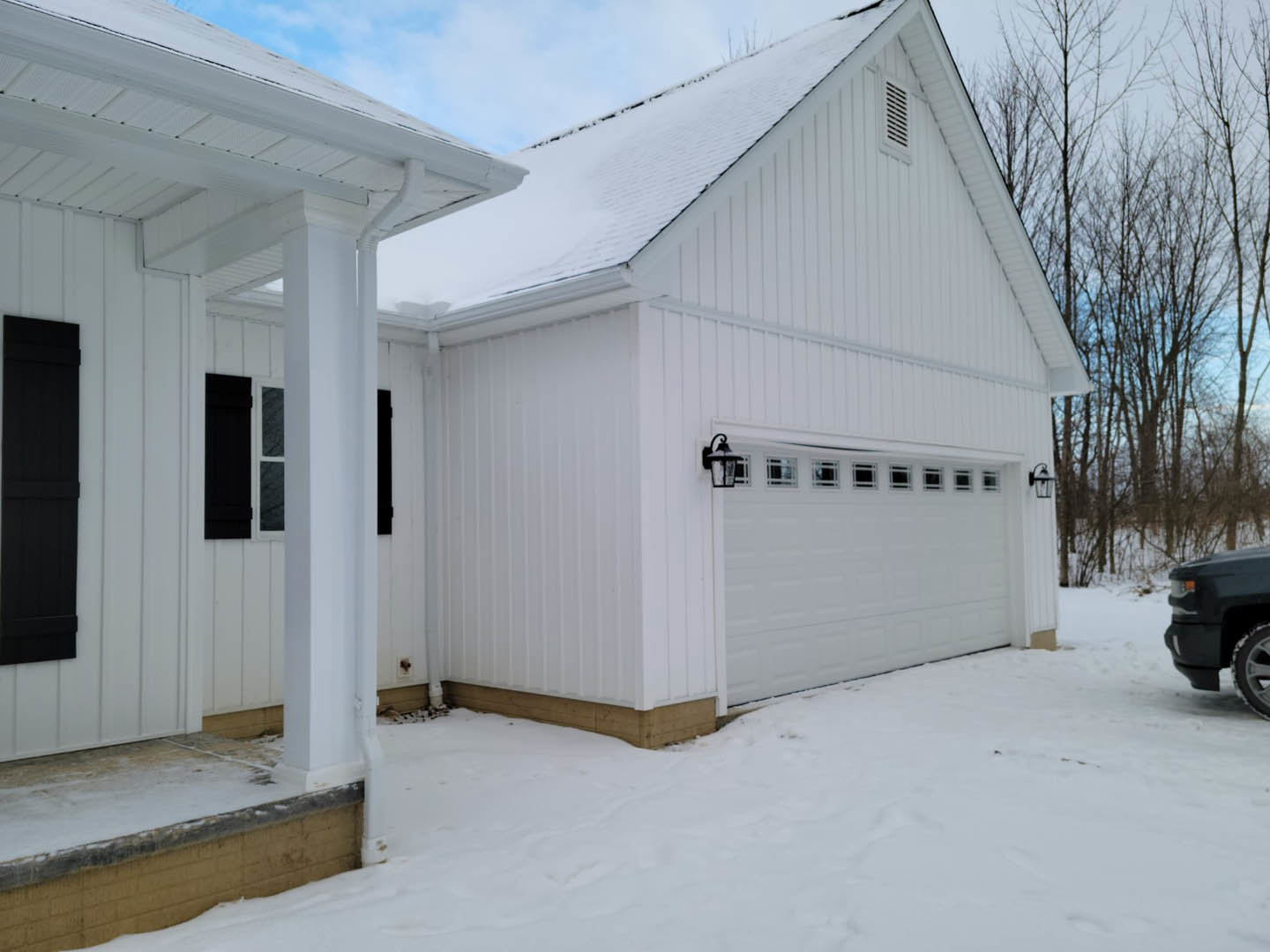 White siding house with attached garage, black shutters, black truck parked in driveway, snow covering ground and roof, leafless trees in background