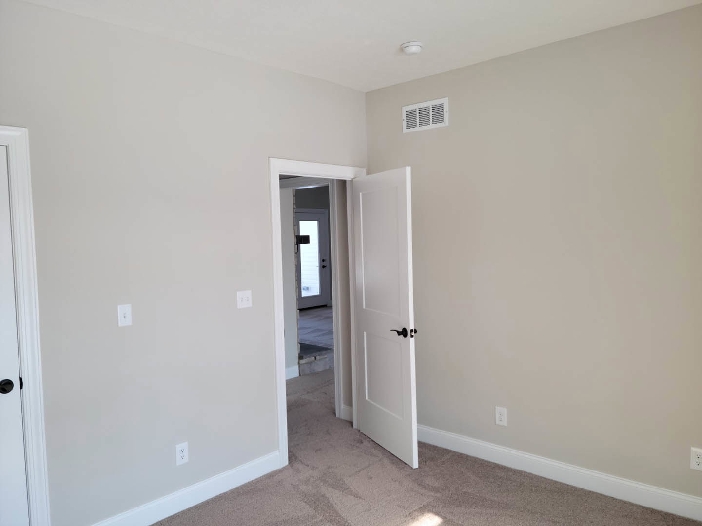 White paneled door with black knob partially open in a room with light plaster walls and wood flooring; wall vent visible nearby
